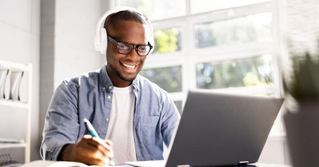 Hombre con audífonos tomando un curso online en su laptop, escribiendo en un cuaderno.